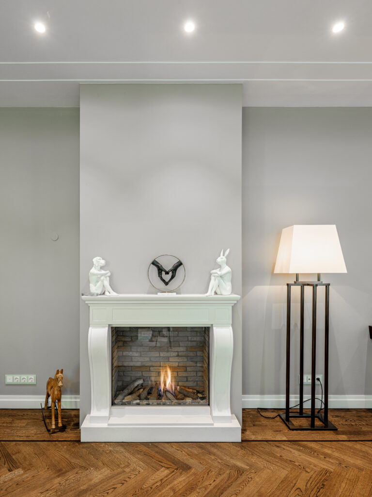 Ein eleganter Kamin mit weißem Rahmen und brennendem Feuer in einem Raum mit Parkettboden. Auf dem Kaminsims stehen zwei weiße Hasenfiguren und eine Skulptur mit zwei Händen, die ein Herz formen. Links neben dem Kamin steht ein hölzernes Schaukelpferd, rechts eine hohe Stehlampe mit weißem Schirm. Die Wände sind hellgrau, und die Decke hat eingebaute Leuchten.
