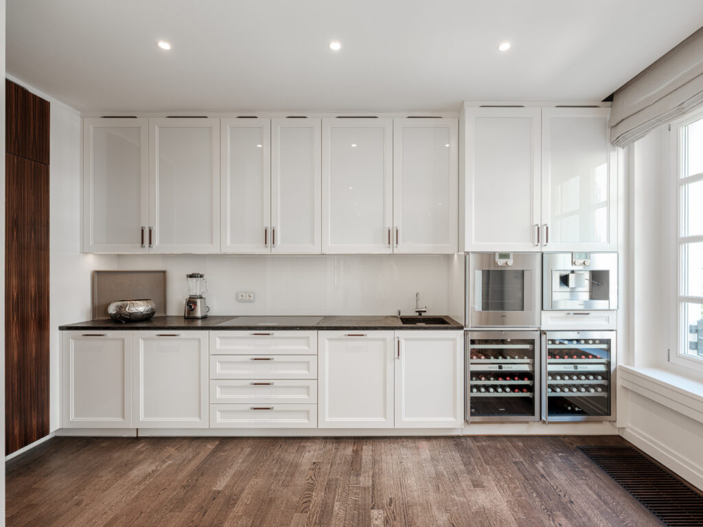 Eine moderne Küche mit weißen Hochglanzschränken und einer dunklen Arbeitsplatte. Auf der Arbeitsplatte stehen ein Mixer und eine Schale, daneben ein Spülbecken und ein Weinkühlschrank mit Glasfront. Der Boden ist aus Holz, und ein großes Fenster mit beigem Vorhang lässt Tageslicht herein. Eingebaute Deckenleuchten sorgen für zusätzliche Helligkeit.