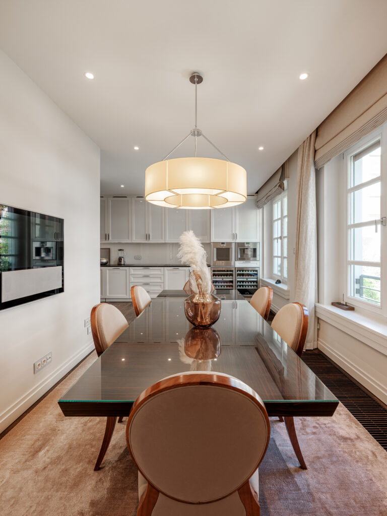 Ein modernes Esszimmer mit einem gläsernen Esstisch, umgeben von vier gepolsterten Stühlen. Über dem Tisch hängt ein großer, runder Kronleuchter. Im Hintergrund ist eine weiße Küche mit Hochglanzschränken, einem Weinkühlschrank und einer Arbeitsplatte mit Küchengeräten zu sehen. Große Fenster mit beige Vorhängen lassen Tageslicht herein, und der Boden ist mit einem Teppich und Parkett ausgelegt.