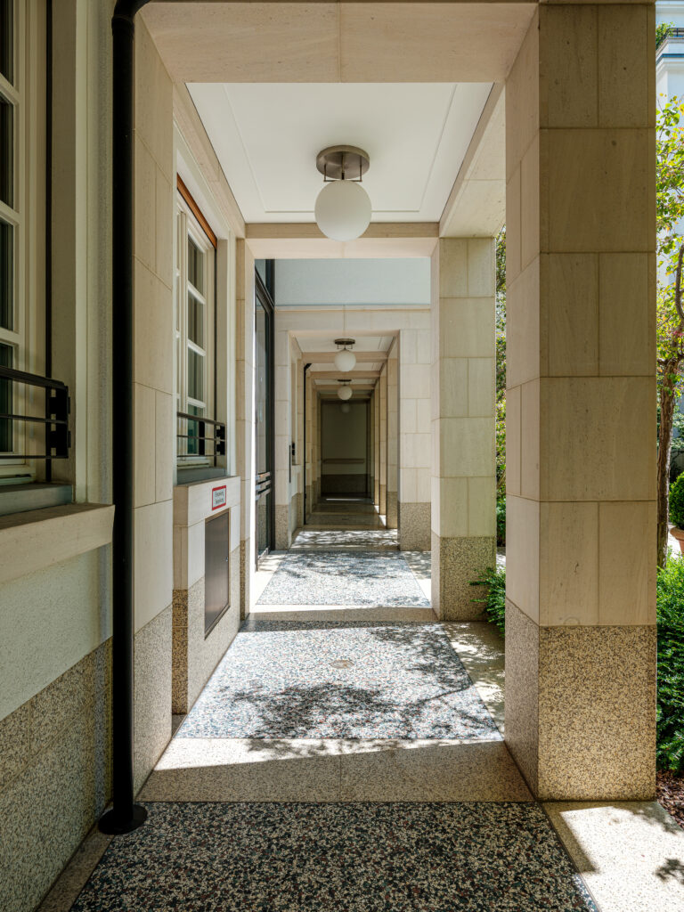 Ein überdachter Laubengang an einem modernen Gebäude mit beigen Steinsäulen und einer Decke mit eingebauten runden Leuchten. Der Boden besteht aus grauem Terrazzo, und Sonnenlicht wirft Schatten von Bäumen auf den Boden. Links ist eine Wand mit einer Fensterreihe, rechts sind Pflanzen und Bäume sichtbar.