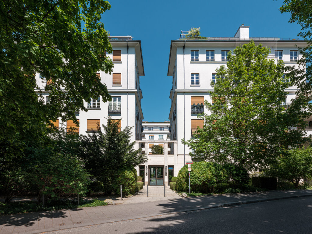 Ein mehrstöckiges Gebäude mit weißer Fassade und braunen Rollläden, umgeben von grünen Bäumen und Sträuchern. Der Eingang ist mittig zwischen zwei Gebäudeteilen, mit einem gepflasterten Weg und einem kleinen Vorgarten. Der Himmel ist klar und blau, und Sonnenlicht wirft Schatten auf den Boden.