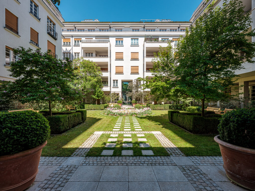 Ein gepflegter Innenhof mit einem symmetrischen Garten, umgeben von einem mehrstöckigen weißen Gebäude mit Holzfensterläden. Ein gepflasterter Weg führt durch das Zentrum des Gartens, gesäumt von grünen Hecken, großen Terrakottatöpfen mit Büschen und schattenspendenden Bäumen. Im Hintergrund sind Blumenbeete und Bänke zu sehen, während die Sonne den Rasen erhellt.
