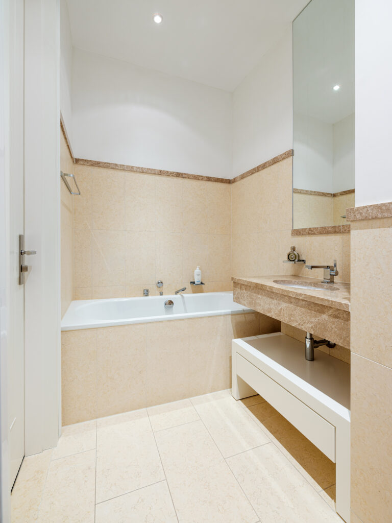 Ein modernes Badezimmer mit beige-weißen Fliesen und einer Marmorarbeitsplatte. Links steht eine weiße Badewanne mit silbernen Armaturen, daneben ein Waschbecken mit einem großen Spiegel darüber. Auf der Arbeitsplatte stehen Pflegeprodukte, und ein Handtuchhalter ist an der Wand befestigt. Die Beleuchtung kommt von eingebauten Deckenleuchten.
