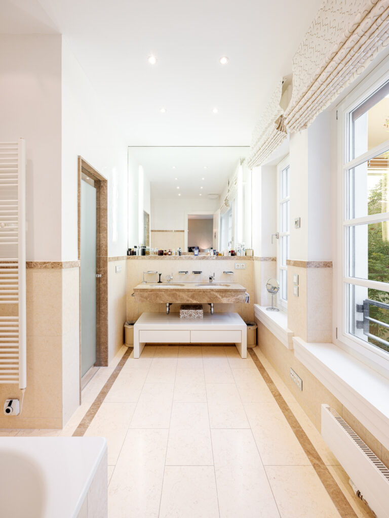 Ein modernes Badezimmer mit beigen Fliesen und einer großen Badewanne links. Rechts steht ein Doppelwaschbecken aus Marmor auf einem weißen Unterschrank, darüber ein großer Spiegel. Die Decke hat eingebaute Spots, und an den Fenstern hängen helle, gemusterte Vorhänge. Ein Handtuchheizkörper ist links an der Wand, und durch die Fenster fällt Tageslicht herein.