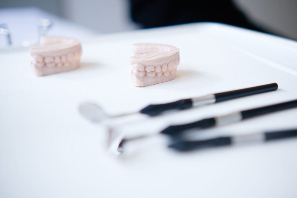 Zwei Zahnmodelle aus Gips, die den Ober- und Unterkiefer darstellen, liegen auf einem weißen Tisch. Im Vordergrund sind zahnmedizinische Instrumente wie ein Mundspiegel und eine Sonde zu sehen. Im Hintergrund ist unscharf ein Zahnarztstuhl erkennbar.