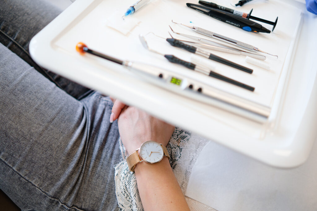 Ein weißes Tablett mit zahnmedizinischen Instrumenten wie Spritzen, Sonden und einem Mundspiegel liegt auf dem Schoß einer Person, die graue Jeans und ein weißes Spitzenoberteil trägt. Die Person trägt eine elegante Uhr mit goldenem Armband am Handgelenk. Im Hintergrund ist ein Teil einer Person in blauem Kittel zu sehen.