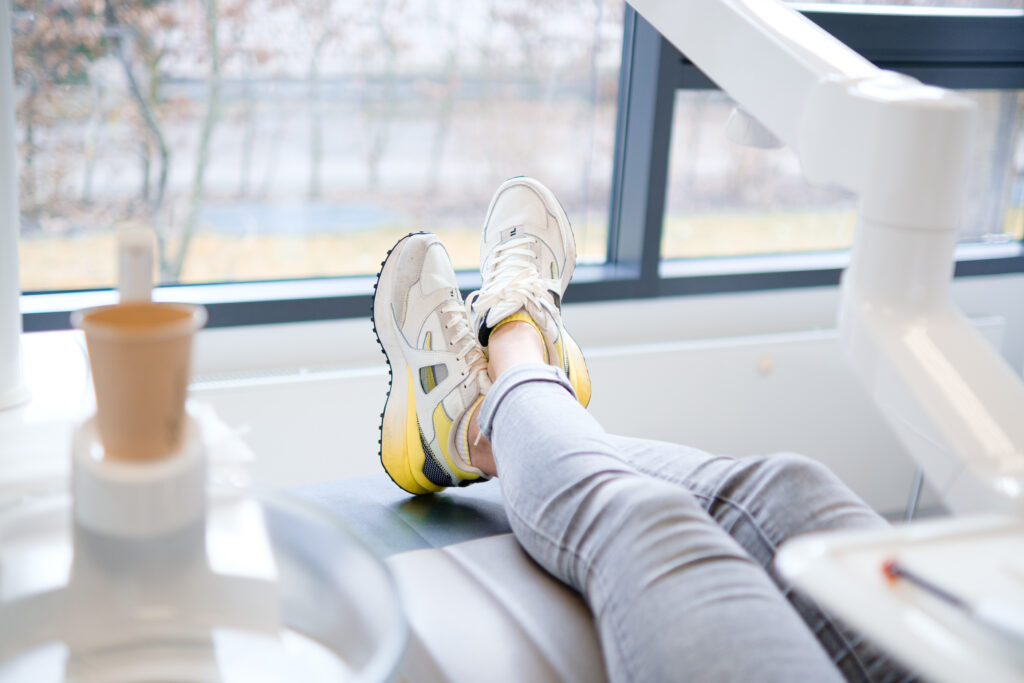 Die Beine einer Person in grauen Jeans und weißen Sneakers mit gelben Akzenten ruhen auf einem Zahnarztstuhl. Neben dem Stuhl steht ein Tablett mit zahnmedizinischen Instrumenten, und im Hintergrund ist ein Fenster mit Blick auf kahle Bäume zu sehen. Ein Becher und ein Teil des Behandlungsraums sind ebenfalls sichtbar.