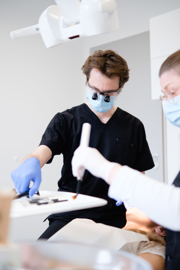 Ein Zahnarzt in schwarzem Kittel und mit blauen Handschuhen sowie einer Lupe auf der Stirn arbeitet an einer Patientin, die auf einem Zahnarztstuhl liegt. Er hält ein zahnmedizinisches Instrument in der Hand. Eine Assistentin in weißem Kittel und mit blauen Handschuhen unterstützt ihn. Auf einem Tablett liegen verschiedene zahnmedizinische Werkzeuge. Der Behandlungsraum ist hell und steril, mit einer Lampe über dem Stuhl.