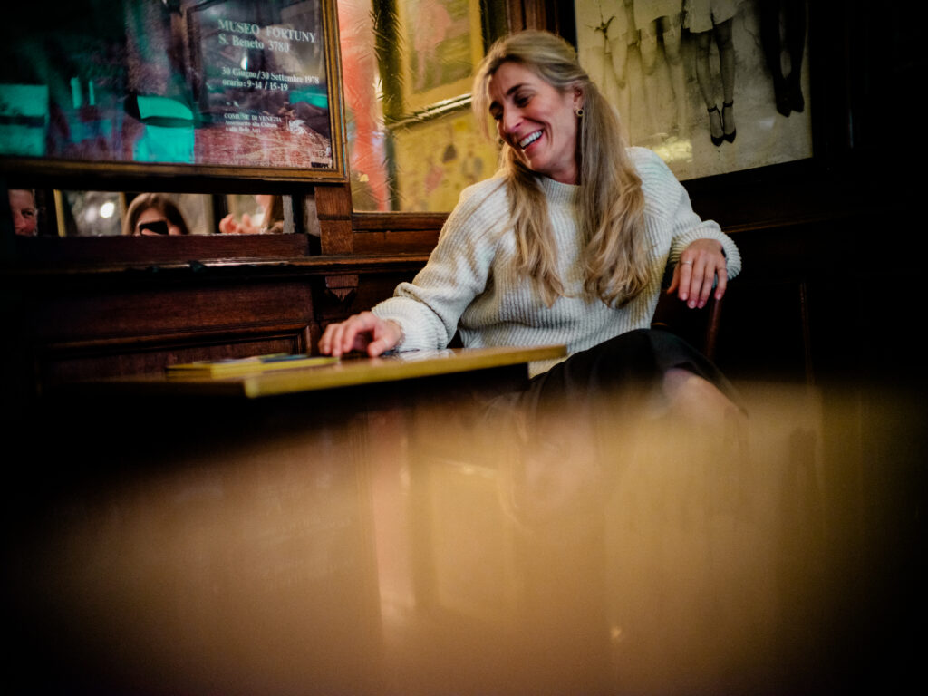 Emotionales Portrait im Orphée Regensburg: Frau im hellen Pullover greift lachend nach der auf dem Tisch liegenden Speisekarte. Im Hintergunrd Wandbilder und Details – Portraitfotografie von Alexander Gerner.