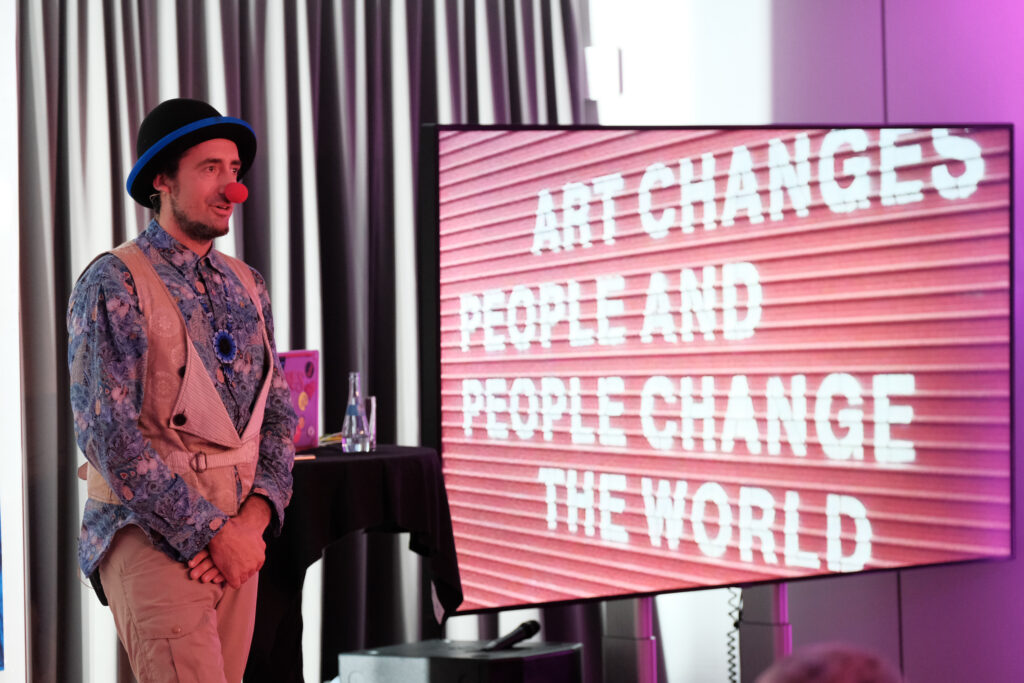 Fabian Seewald bei seinem Vortrag auf dem #nxtKnowledge mit Clownsnase und Bildschirm im Hintergrund mit dem Text „ART CHANGES PEOPLE AND PEOPLE CHANGE THE WORLD“
