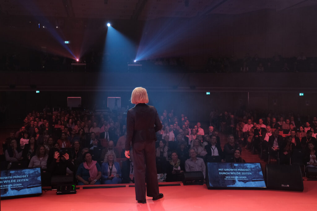 Vaya Wieser-Weber auf der Bühne, fotografiert von hinten mit Blick ins Publikum, Eventfotograf Stuttgart #nxtKNOWLEDGE 2025 Böblingen.