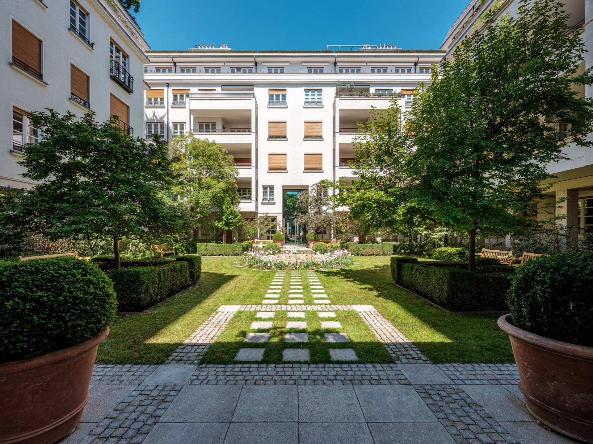 Ein gepflegter Innenhof mit einem symmetrischen Garten, umgeben von einem mehrstöckigen weißen Gebäude mit Holzfensterläden. Ein gepflasterter Weg führt durch das Zentrum des Gartens, gesäumt von grünen Hecken, großen Terrakottatöpfen mit Büschen und schattenspendenden Bäumen. Im Hintergrund sind Blumenbeete und Bänke zu sehen, während die Sonne den Rasen erhellt.